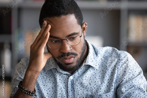 Close up African businessman think search problem solution feels concerned anxious. Guy student in glasses sitting indoors learns educational material, preparing for exams, do task brain work concept