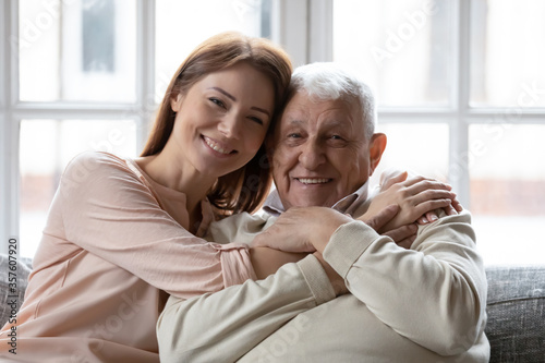 Head shot portrait smiling older father and grownup daughter looking at camera, sitting on cozy couch at home, overjoyed young woman and mature man hugging, cuddling, two generations concept