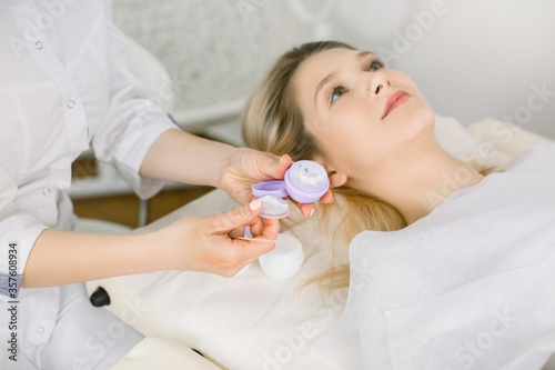 Visiting of beautician. Gorgeous young woman lying on the couch and waiting for relax and beauty procedures in spa center. Hands of female doctor holding bottle of cream. Focus on hands with cream