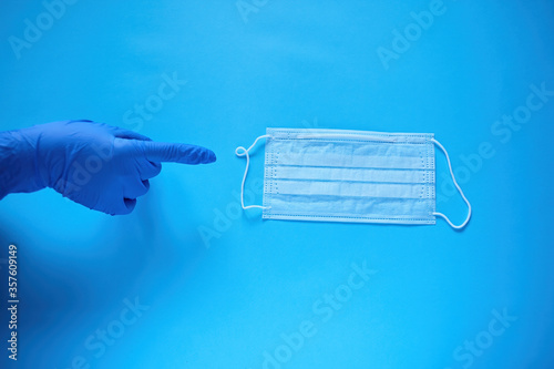A hand in a protective medical glove points a finger at a protective medical mask. Protect from coronavirus concept on blue background