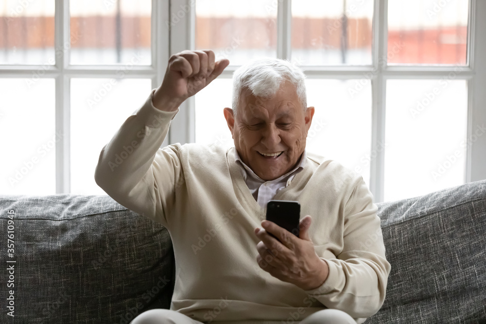 Overjoyed smiling mature man reading good news in smartphone, looking at phone screen, showing yes gesture, sitting on couch, excited happy elderly male celebrating success, online lottery win