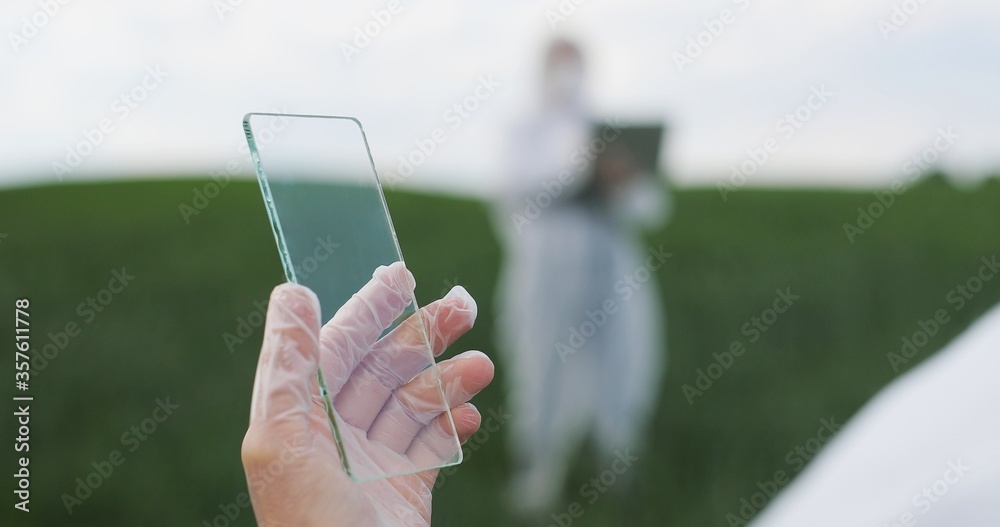 View over shoulder of farmer ecologist in protective suit tapping on glass transparent screen. Close up of futuristic device in field Touchscreen of hi-tech Scientist with smartphone of future in hand