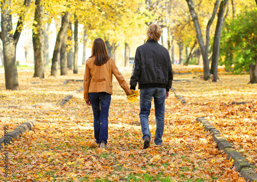 A couple in love, a guy and a girl holding hands goes through the autumn Park. Rear view. A bright autumn day, lots of fallen leaves under your feet.