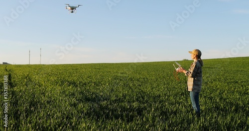 Caucasian woman farmer in hat standing in green wheat field and controlling of drone which flying above margin. Female using tablet device as controller. Technologies in farming. Rear. Back view.
