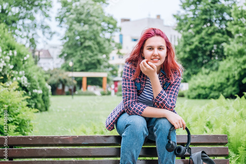 Portrait of Beautiful modern young female teenager with extraordinary hairstyle color in a checkered shirt with wireless headphones on the neck sitting on the city park bench and smiling at the camera