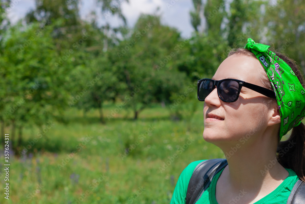 Portrait of a pretty hipster girl in a sunglasses, green shirt and bandana standing on a nature background on sunny day. Positive emotional caucasian young long haired lady smiles and looks on the sky Portrait of a pretty hipster girl in a sunglasses, green shirt and bandana standing on a nature background on sunny day. Positive emotional caucasian young long haired lady smiles and looks on the sky