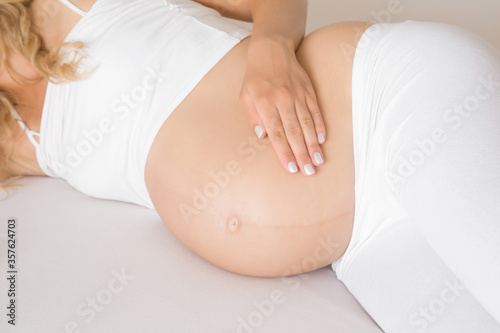 Young woman in white clothes lying down and feeling her baby move. Hand touching naked belly. Emotional loving pregnancy time - 35 weeks. Baby expectation. Love, happiness concept. Closeup. 9 month.