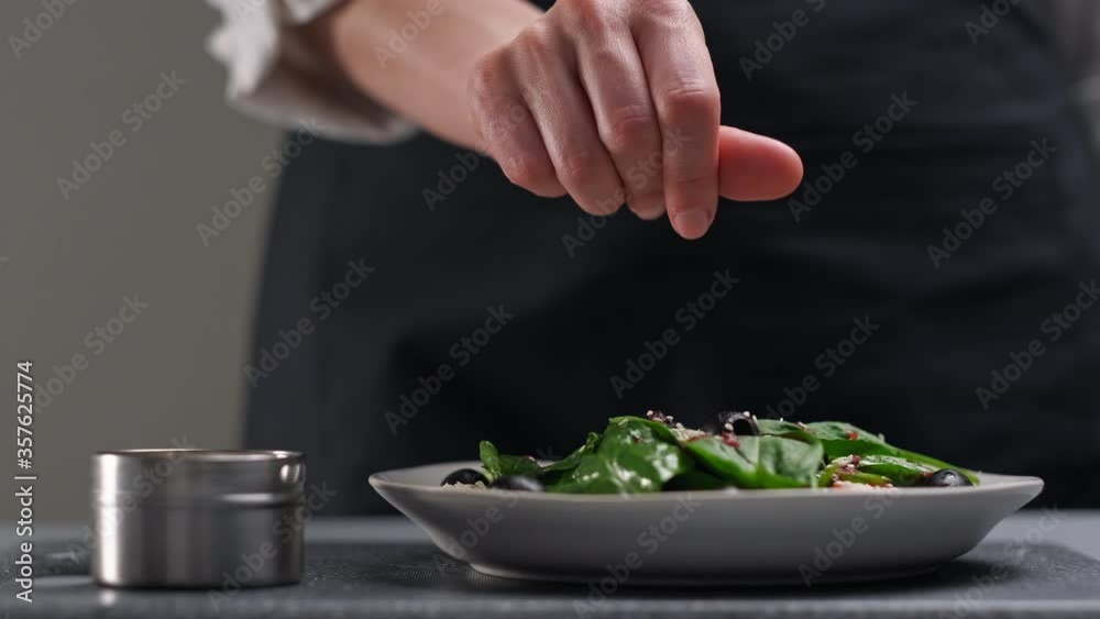 A female chef in a white uniform and a black apron in the restaurant kitchen. Cooking. The cook sprinkles the salad with sesame seeds. A female chef in a white uniform and a black apron in the restaurant kitchen. Cooking. The cook sprinkles the salad with sesame seeds.