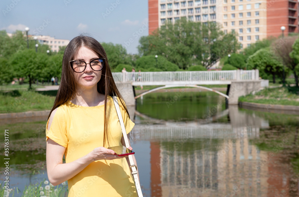 Obraz premium young woman with long hair in a yellow dress with a phone stands in the background of a building on the Bank of a pond