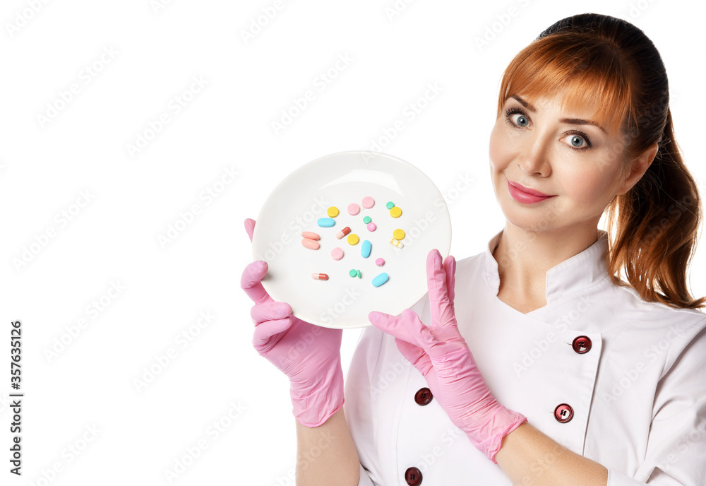Portrait of young beautiful red-haired woman doctor or nurse in white special uniform and gloves showing plate with colorful pills Portrait of young beautiful red-haired woman doctor or nurse in white special uniform and gloves showing plate with colorful pills