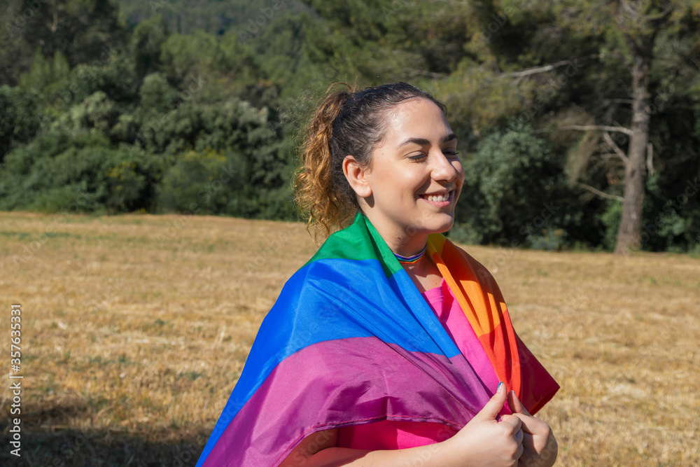 Young woman with the rainbow flag around her shoulders smiling on outdoors. Lgtbiq concept. Young woman with the rainbow flag around her shoulders smiling on outdoors. Lgtbiq concept.
