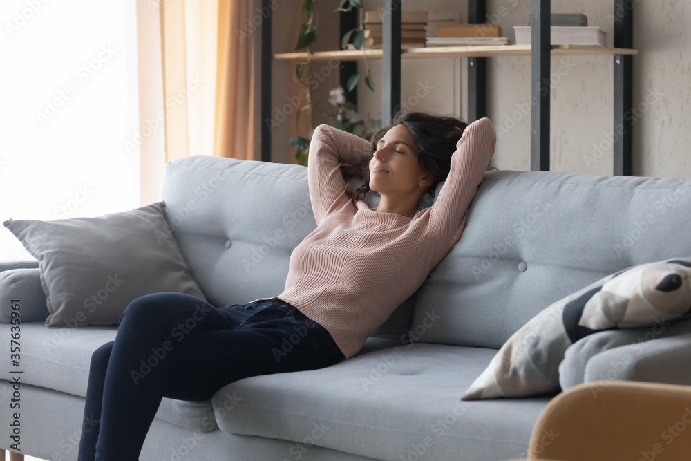 Relaxed satisfied young woman resting on cozy couch at home, calm beautiful  girl leaning back with hands behind head, enjoying lazy weekend,  daydreaming, sitting on sofa in living room Stock Photo