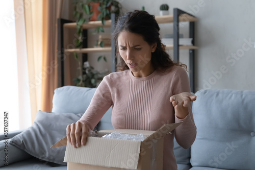 Angry confused woman unpacking parcel, wrong or broken online store order, sitting on couch at home, dissatisfied female looking in cardboard box, bad delivery service, displeased by post shipping photo