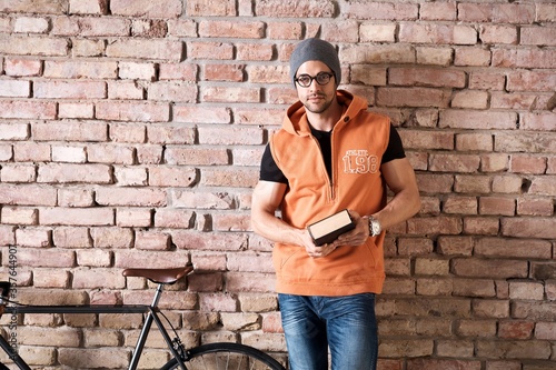 Young smart trendy caucasian man in glasses and cap standing at brick wall reading book. Copyspace for text. " Zima Adam
