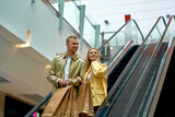 young beautiful married couple on escalator in shopping mall, caucasian man and woman in coats holding bags after shopping