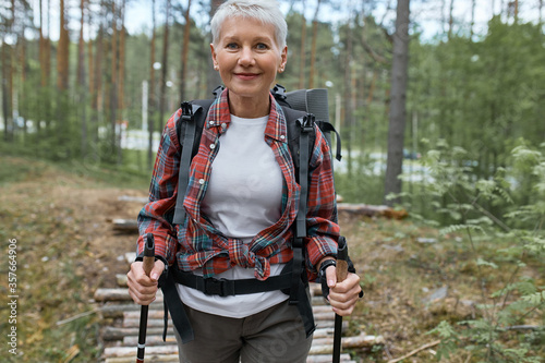 Outdoor activities, people and vacations concept. Attractive short haired middle aged woman in activewear hiking in forest using poles for nordic walking, doing aerobic workout, enjoying nature