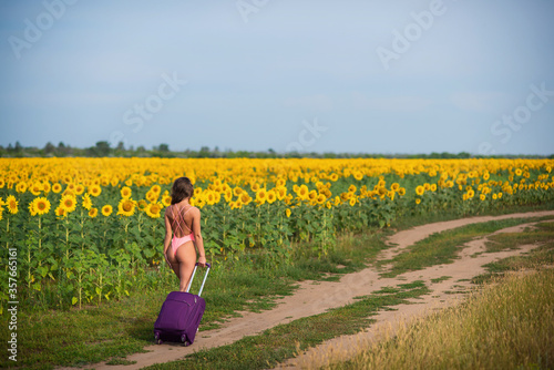 Beautiful woman in a pink swimsuit travels through the countryside with a suitcase. A girl in a monokini is walking alone on a field of sunflowers with a big bag. Hiking. Rest and social distance.