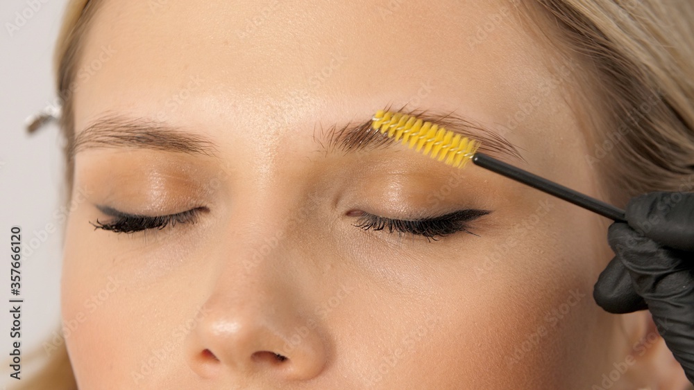 Stylist does professional makeup for beautiful woman at the beauty salon. Gloved master combing patient’s eyebrows with a special brush. Eyebrow shaping with a cosmetic brush close-up. 