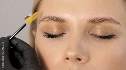 Stylist does professional makeup for beautiful woman at the beauty salon. Gloved master combing patient’s eyebrows with a special brush. Eyebrow shaping with a cosmetic brush close-up. 