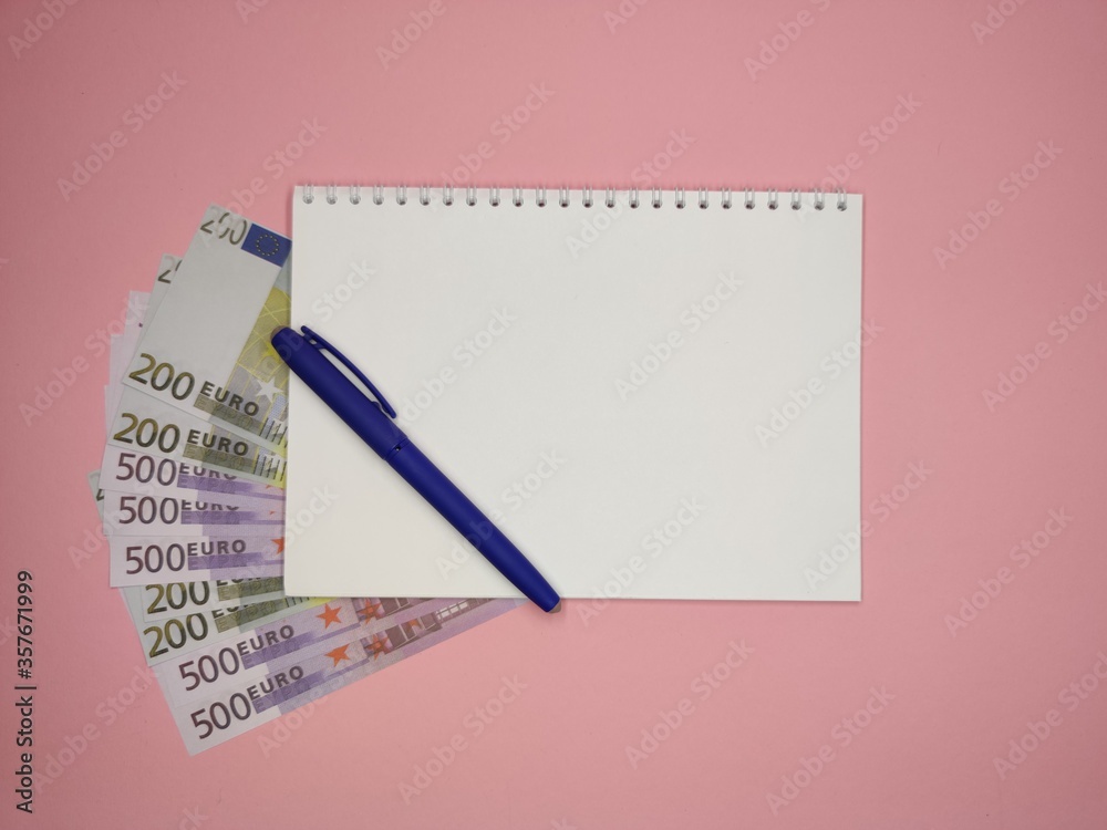 on a pink background, a blue pen on the left diagonally on a Notepad with a copy space, 200 and 500 Euro bills peek out from under it. on a pink background, a blue pen on the left diagonally on a Notepad with a copy space, 200 and 500 Euro bills peek out from under it.