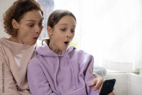 Stunned Caucasian mom and teen daughter feel surprised reading online news on cellphone at home, amazed mother and teenage girl child shocked by internet offer or deal, shopping on smartphone