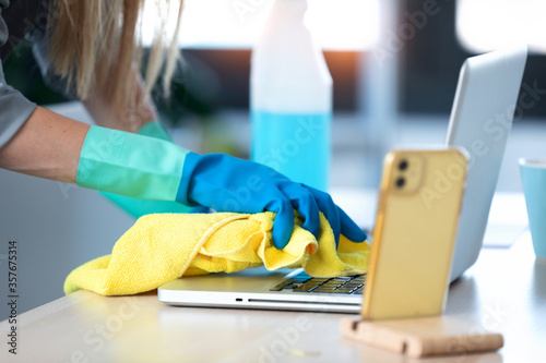 Woman with protective gloves cleaning the laptop with spray and rag in office.