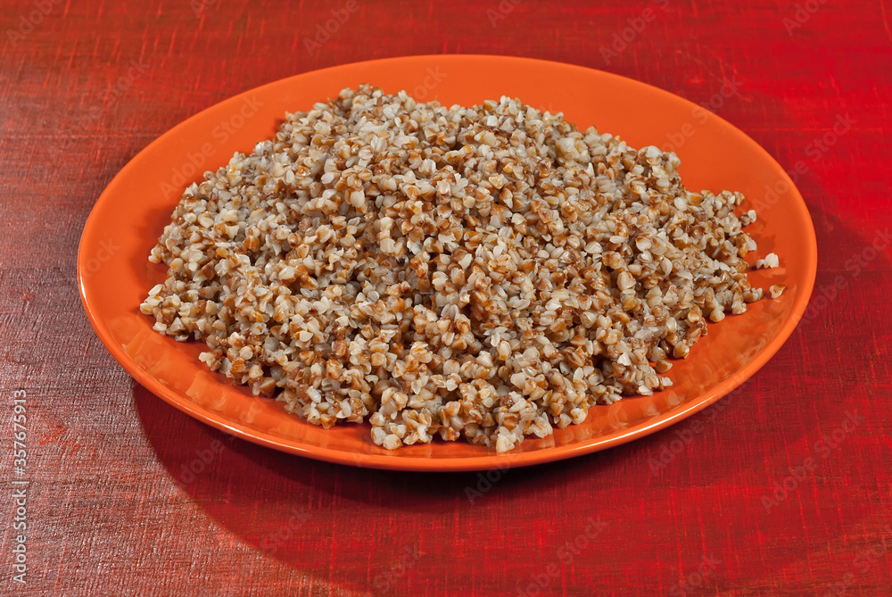 Buckwheat orange plate on a wooden table. Food in utensils on a background of old red plywood. Buckwheat orange plate on a wooden table. Food in utensils on a background of old red plywood.