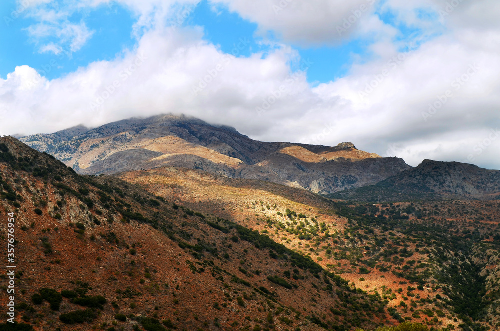 View on mountains with low hanging clouds and blue sky and green trees. Mountains and shadows landscape. Tourism in Europe concept. Autumn in Crete