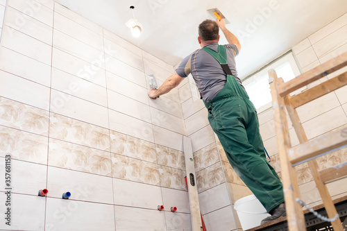 A construction worker applies leveling compound to the ceiling with a stainless steel trowel. A builder dressed in protective clothing is standing on a ladder. © fotodrobik