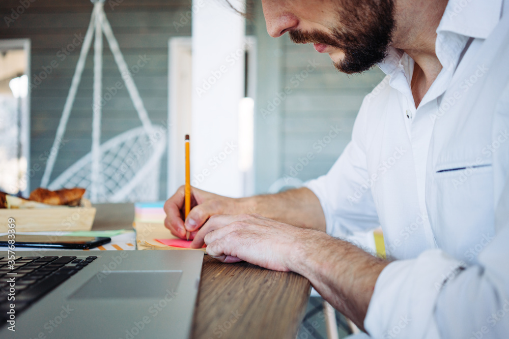 Young man with stubble working at home in bright wooden room in his own house. Remote workplace. Writing with pencil on a sticker Young man with stubble working at home in bright wooden room in his own house. Remote workplace. Writing with pencil on a sticker
