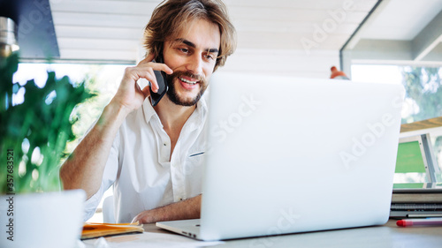 Young man with stubble working at home in bright wooden room in his own house. Remote workplace. Smiling and talking on the phone