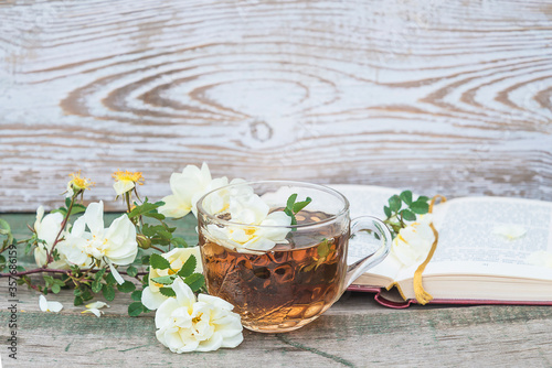 Tea time concept. Glass cup of tea, twig with white wildroses and open book on rustic wooden background. Space for text photo