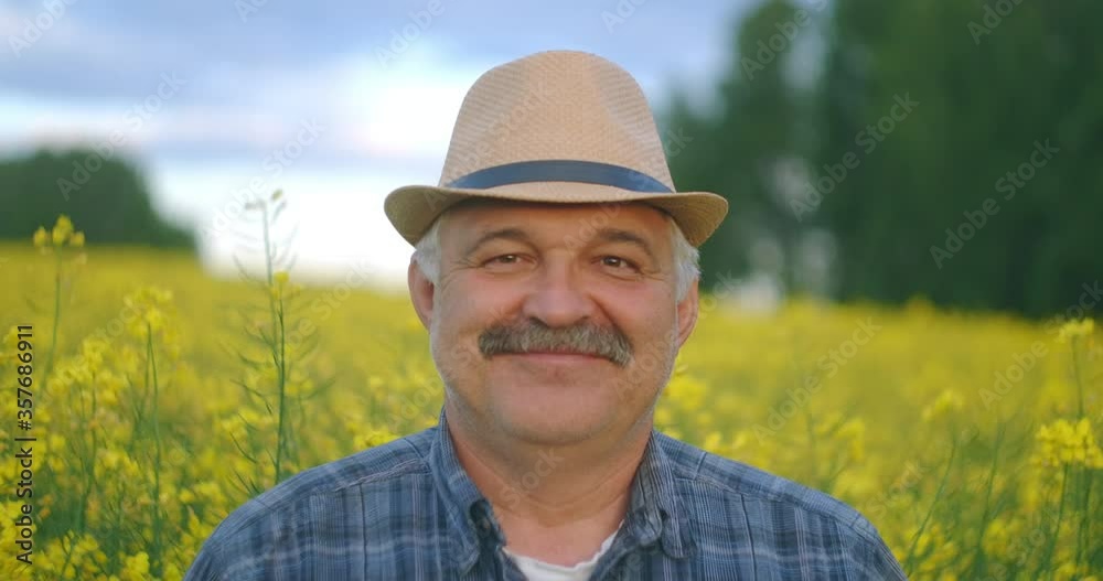 Close up of the senior Caucasian Looking at the camera good looking wise man farmer in a hat looking at the side, turning face to the camera and smiling in the wheat rapeseed field. Portrait Close up of the senior Caucasian Looking at the camera good looking wise man farmer in a hat looking at the side, turning face to the camera and smiling in the wheat rapeseed field. Portrait