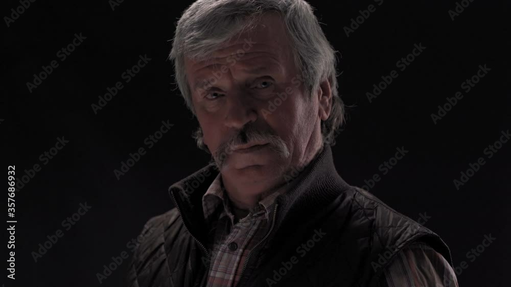 Grey haired old man thoughtful looking down then on at camera and down with camera slide on wearing jacket in semi lit light studio isolated on black background. Prores 422. Grey haired old man thoughtful looking down then on at camera and down with camera slide on wearing jacket in semi lit light studio isolated on black background. Prores 422.