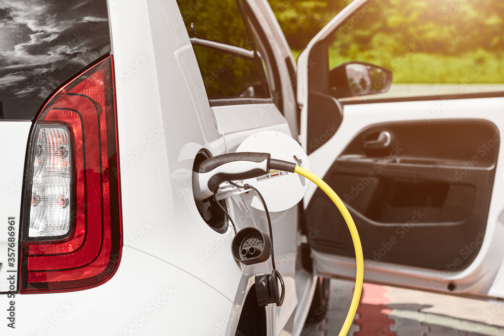 Naklejka premium Yellow electric socket charging an electric car in the street at sunlight. Power supply plugged into the electric automobile