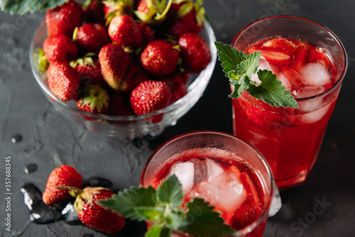 Closeup refreshing summer drink of strawberries and mint, juice, soda with ice cubes and berry slices, in a beautiful glass with water droplets on a wooden board, dark background