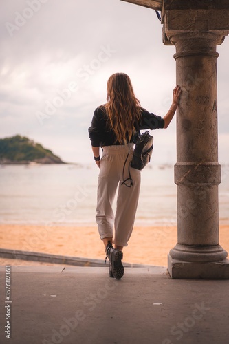 Lifestyle in the city with a blonde girl in white pants and a leather jacket near the beach. Photos next to a column on the beach