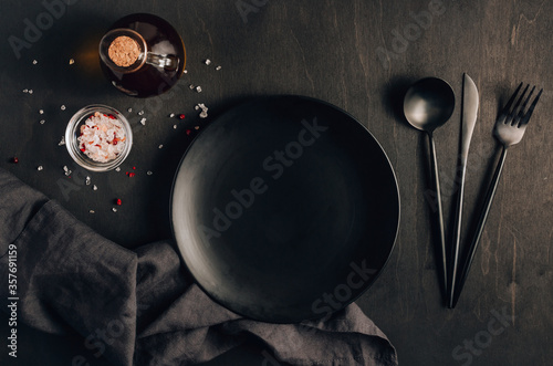 Black table setting: plate, napkin, silverware, olive oil and salt on black wooden background