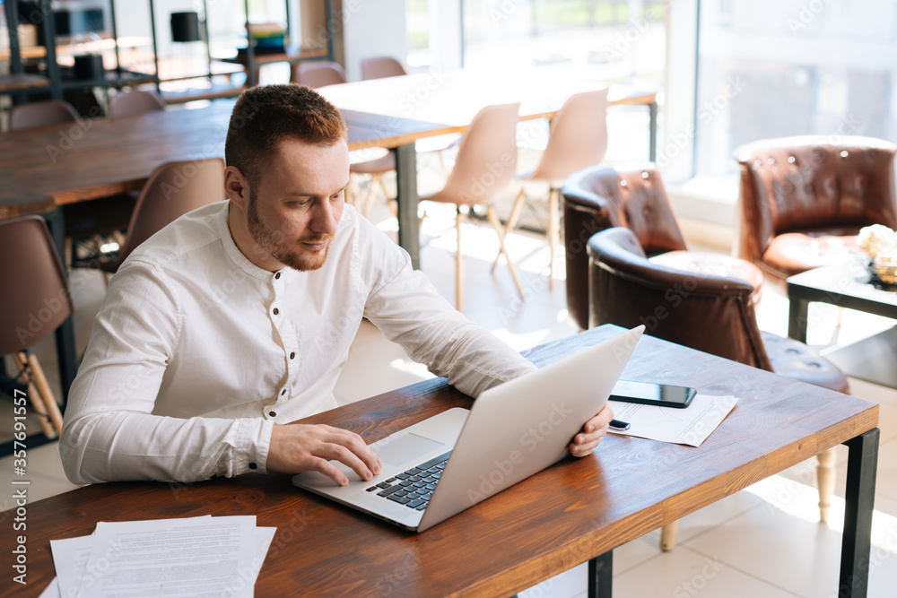 Top view of happy young man wearing fashion casual clothing is working on laptop computer at the table in modern office room on background of large window. Concept of office working. Top view of happy young man wearing fashion casual clothing is working on laptop computer at the table in modern office room on background of large window. Concept of office working.