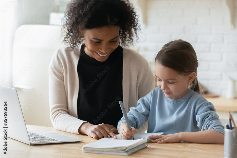 African tutor engaged with schoolgirl help complete tasks seated at table at home. Concept of education kid development, european child homeschooling writing homework with american stepmother concept