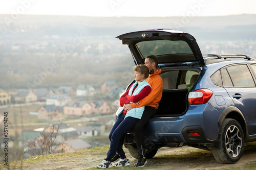 Happy couple standing together near a car with open trunk enjoying view of rural landscape nature. Man and woman leaning on family vehicle luggage compartment. Weekend travel and holidays concept. © bilanol