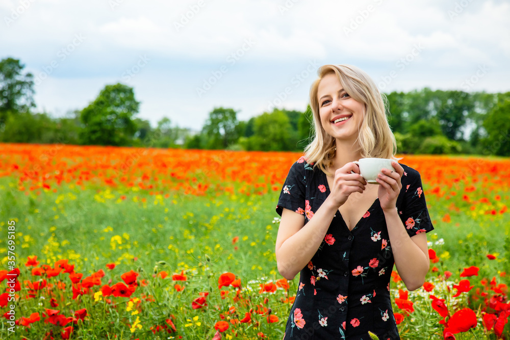 Blonde girl in beautiful dress with cup of coffee in poppies field in summer time