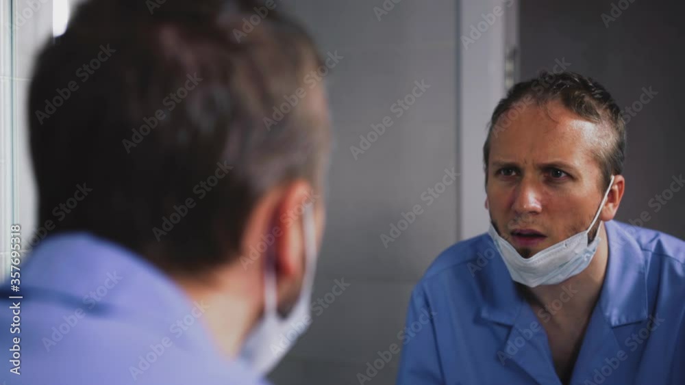 close-up of young uncut bearded doctor, dressed in surgical care suit, in front of mirror, takes off his glasses, protective mask, scared, frowns, looks to side, ponders