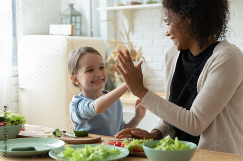 Caucasian daughter give high five gesture to african mom cooking together veggie salad in kitchen, chopped fresh ingredients in bowls on dining table. Eat healthy homemade food, family recipe concept