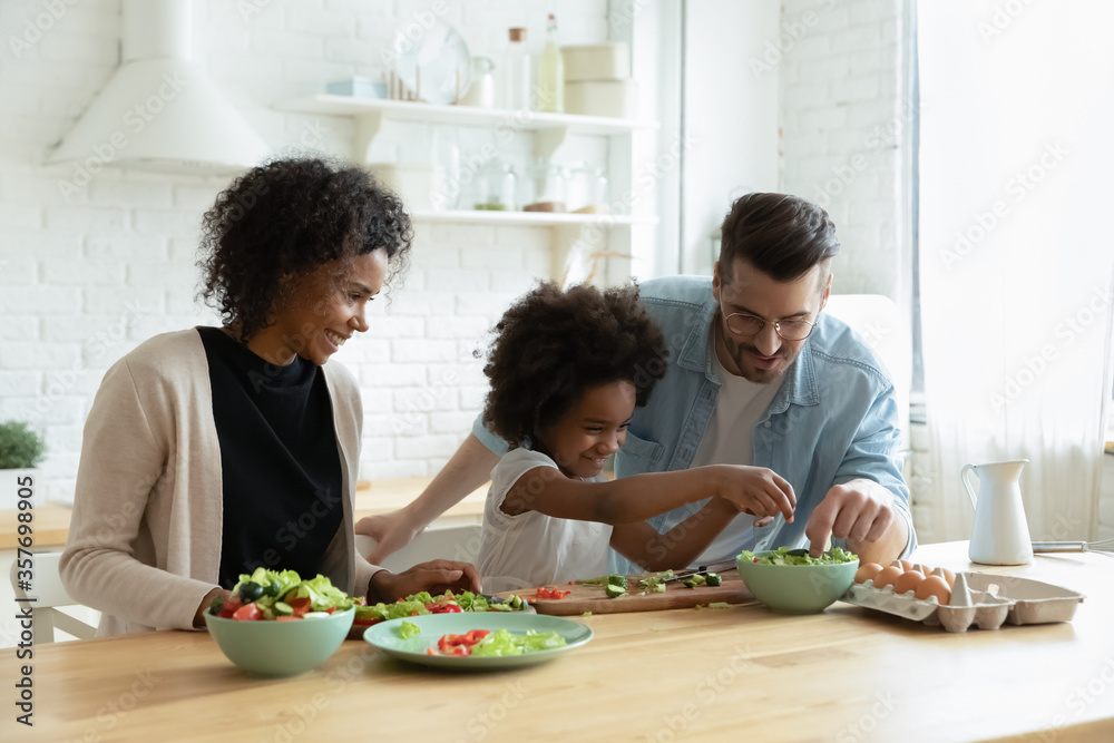 Caring multiethnic couple teach small daughter cook vegetable salad in modern domestic kitchen. Family enjoy cooking and communication together, healthy organic food preparation, love and bond concept Caring multiethnic couple teach small daughter cook vegetable salad in modern domestic kitchen. Family enjoy cooking and communication together, healthy organic food preparation, love and bond concept