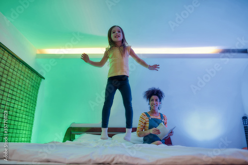 A nanny for fun learning. African american woman baby sitter and cute little girl reading book in the evening. Nanny holding book while kid jumping on the bed, enjoying listening to fairy tale