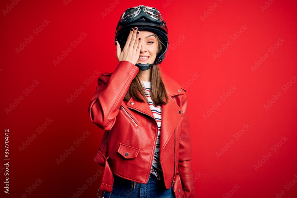 Young beautiful brunette motocyclist woman wearing motorcycle helmet and red jacket covering one eye with hand, confident smile on face and surprise emotion.