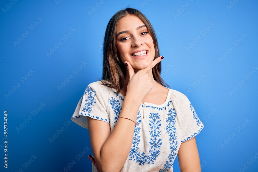 Young beautiful brunette woman wearing casual t-shirt standing over blue background looking confident at the camera smiling with crossed arms and hand raised on chin. Thinking positive. Young beautiful brunette woman wearing casual t-shirt standing over blue background looking confident at the camera smiling with crossed arms and hand raised on chin. Thinking positive.