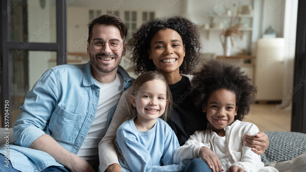 Multinational family married couple and daughters sit on couch looks at camera photo shooting feels happy. Cute multi-ethnic girls and parents portrait, homeowners, new home, prosperous future concept