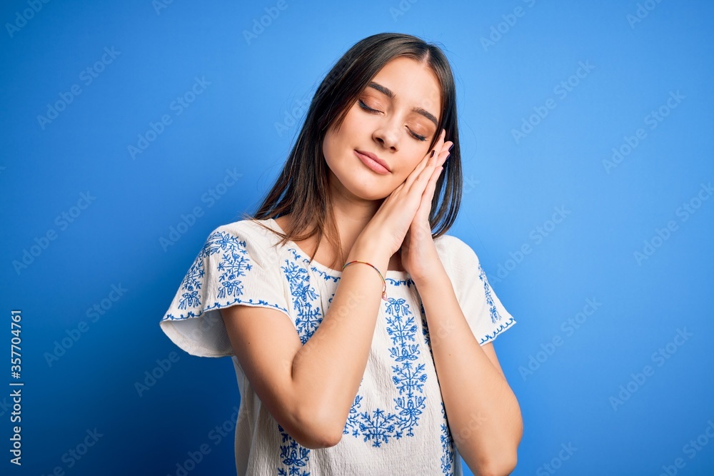 Young beautiful brunette woman wearing casual t-shirt standing over blue background sleeping tired dreaming and posing with hands together while smiling with closed eyes.